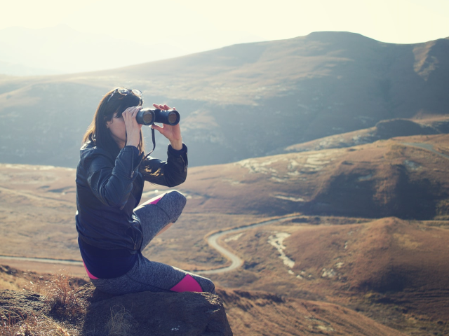 Woman sitting on mountain ridge looking through binoculars